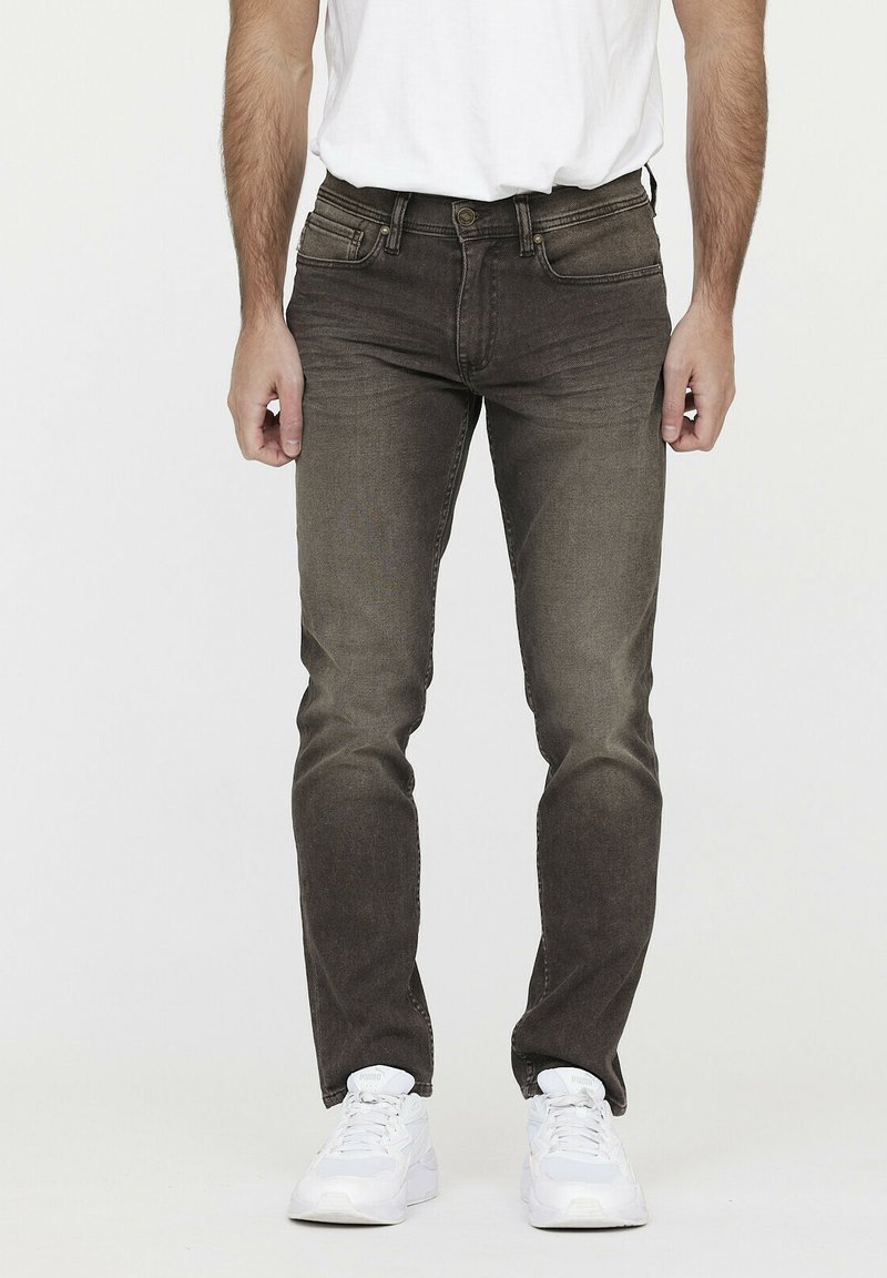 Jean foncé en marron avec une coupe slim, légères décolorations, cinq poches, fermeture éclair, et coutures contrastantes, associés à des baskets blanches.