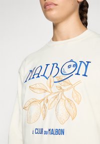 Krémová mikina s modrou a zlatou výšivkou. Design obsahuje text "MALBON" a grafiku citronové větvičky. Měkká textura, kulatý výstřih.