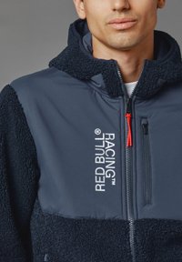 Fleece- en nylonjack in marineblauw, met een gestructureerd bovenstuk, Red Bull Racing-logo en een ritszak met rode accenten.