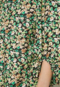 Tissu noir avec motif floral vert, blanc et marron, froncé au niveau du poignet de la manche, laissant apparaître la peau de l’avant-bras.