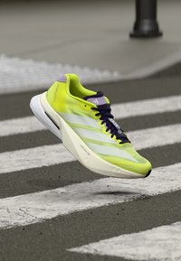 Chaussure de course vert citron avec tige en mesh respirant, rayures blanches et accents violets. Dispose d'une semelle blanche épaisse pour l'amorti.