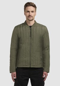 Oliven grøn quiltet bomberjakke med ribbet krave, frontlynlås og sidelommer. Stoffet er glat med en mat finish.