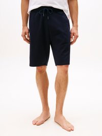 Uomo vestito con pantaloni corti blu navy a cordoncino e una maglietta bianca, in piedi a piedi nudi su uno sfondo bianco.