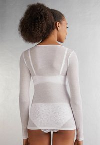 Long-sleeve white ribbed top featuring a scoop back design. Lace detailing can be seen at the lower hem, revealing underneath garments.