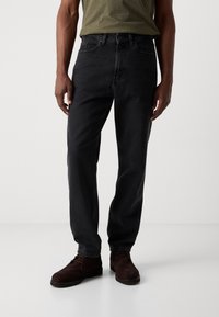 Black denim jeans with a straight leg fit, featuring five pockets and a button closure, paired with brown suede shoes.