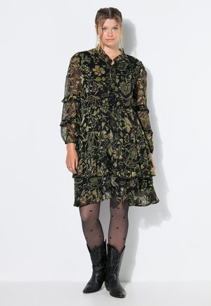 Mujer que lleva un vestido negro transparente con estampado floral y mangas largas, medias transparentes con estampado de corazones y botas de vaquero negras, de pie frente a un fondo blanco.