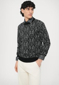 Maglione nero con motivo logo grigio, collo e polsini a costine e dettagli testurizzati. Indossato con pantaloni chiari.