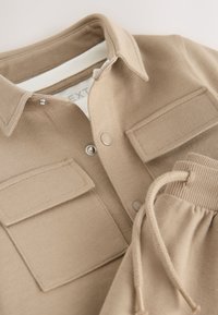 Chemise beige à col avec deux poches poitrine, boutons et détail assorti aux poignets. Fabriquée en tissu doux à texture lisse.