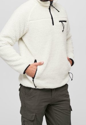 Mann trägt weißen Fleecepullover mit schwarzen Reißverschlussdetails und dunkle Cargohosen, Hände in den Taschen, vor einfarbigem Hintergrund.