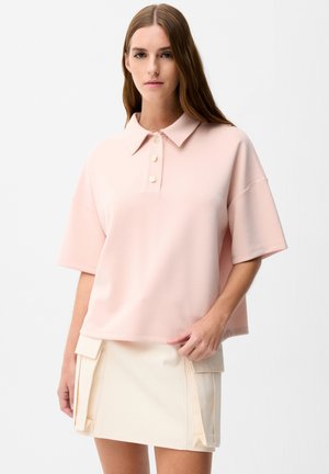 Femme aux longs cheveux bruns portant une chemise rose clair à manches courtes avec col et une jupe couleur crème avec des poches à l'avant.