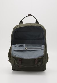Sac à dos en tissu vert avec compartiment principal ouvert montrant des poches intérieures zippées et en mesh, détails en sangle de cuir marron, sur fond blanc.