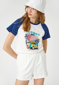 Vit och blå t-shirt med kort ärm som har en färgglad grafik av Malibu Beach. Kombinerad med shorts med elastisk midja i vitt.