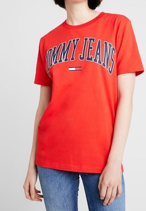 Red short-sleeve T-shirt with "TOMMY JEANS" text and logo, worn by a person in blue jeans, shown from neck to hips.