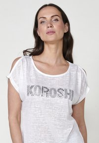 Bílé tričko s krátkým rukávem, volného střihu a výřezem na rameni. Obsahuje černý text "KOROSHI" v stylizovaných vzorech, texturovaná tkanina.