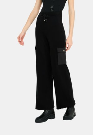 Kobieta ubrana w czarne spodnie o szerokich nogawkach z boczną kieszenią na ładunek oraz czarne botki, stojąca na jednolitym tle.