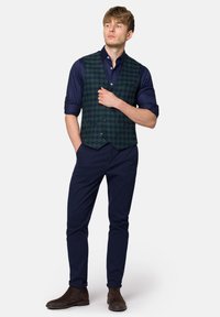 Zielona i granatowa kamizelka w kratę, noszona na ciemnej koszuli, w zestawieniu z ciemnoniebieskimi spodniami i brązowymi botkami za kostkę. Starannie dopasowana, nowoczesny krój.