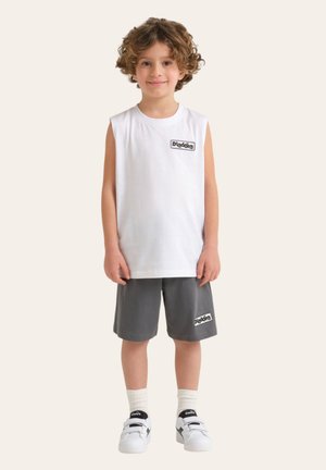 Bambino piccolo con capelli ricci che indossa una maglietta bianca senza maniche e pantaloncini grigi, entrambi con il logo "Diadora", calzini bianchi e scarpe da ginnastica bianche.