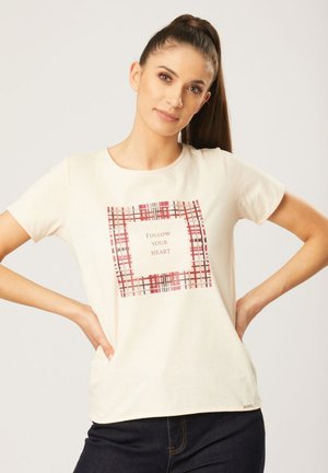 T-shirt couleur crème avec un motif graphique carré présentant un motif écossais rouge et noir et le texte "Suivez votre cœur" au centre.