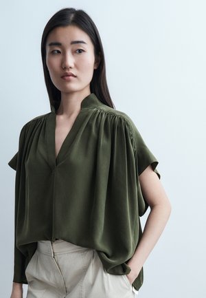 Mlada ženska z dolgimi temnimi lasmi nosi ohlapno olivno zeleno bluzo in svetlo bež hlače, stoji ob enobarvnem svetlo sivem ozadju.