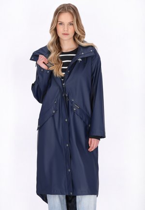 Tânără cu păr blond lung, purtând o haină de ploaie albastru închis peste o cămașă cu dungi alb-negru, ținând gulerul cu o mână.