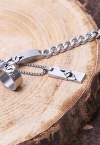Metalene sieradenset met een kettingarmband, een hanger aan een bolletjes ketting en een ring. Alle stukken zijn zilverachtig en hebben een in elkaar grijpend patroon.