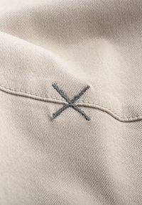 Beige stof met een fijne textuur, voorzien van een grijze geborduurde 'X' en een naad die erlangs loopt. Focus op de stikdetails.