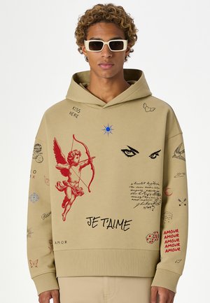 Junge Person mit lockigem Haar trägt einen beigen Hoodie, verziert mit rotem Amor, schwarzen Augen und Text, der "JE T'AIME" und "AMOUR" wiederholt.