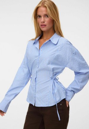 Femme blonde portant une chemise bleu et blanc rayée à boutons avec des liens latéraux, associée à un pantalon marron foncé, posant devant un fond uni.