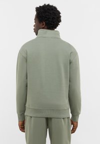 Mężczyzna stoi plecami, ubrany w jasnozieloną bluzę z wysokim kołnierzem i pasujące spodnie, na tle jednolitego białego tła.