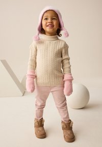 Pull en tricot beige, moufles roses et pantalon, avec des bottes marron ornées de fourrure et de broderies. Un chapeau à pompon rose complète la tenue.