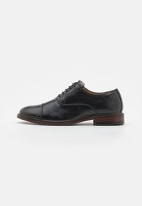 Chaussure de ville en cuir noir avec un bout arrondi, lacée, à texture subtile et à talon en bois brun. Silhouette classique et élégante.
