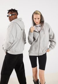 Grå hettegenser med glidelås og løs passform, med en stor hette og sidelommer; vist sammen med sorte bukser og leggings på modellene.