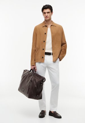 Mężczyzna w beżowej kurtce, białej koszuli, białych spodniach, czarnym pasku i mokasynach, trzymający w prawej ręce dużą ciemnobrązową skórzaną torbę.
