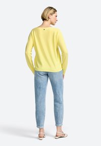 Gelbes, langärmliges Sweater mit weichem Material, weitem Ausschnitt und einem kleinen Akzent am Rücken. Kombiniert mit hellblauen Jeans.