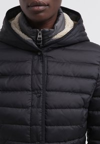 Veste matelassée noire avec un col zippé et une fermeture à boutons. Elle présente une doublure douce, un motif quilté horizontal et une capuche.