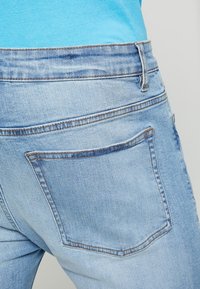 Jeans in denim azzurro chiaro con una singola tasca posteriore, cuciture visibili e una texture liscia, mostrati da una prospettiva posteriore.