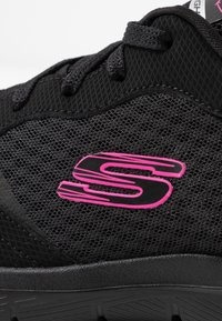 Scarpa atletica nera con tomaia in tessuto a rete, dettagli testurizzati e un logo rosa prominente. Presenta lacci neri e una suola in gomma.