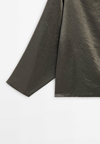 Tissu vert olive foncé froissé à manches longues avec une texture légèrement brillante, présenté sur un fond blanc.