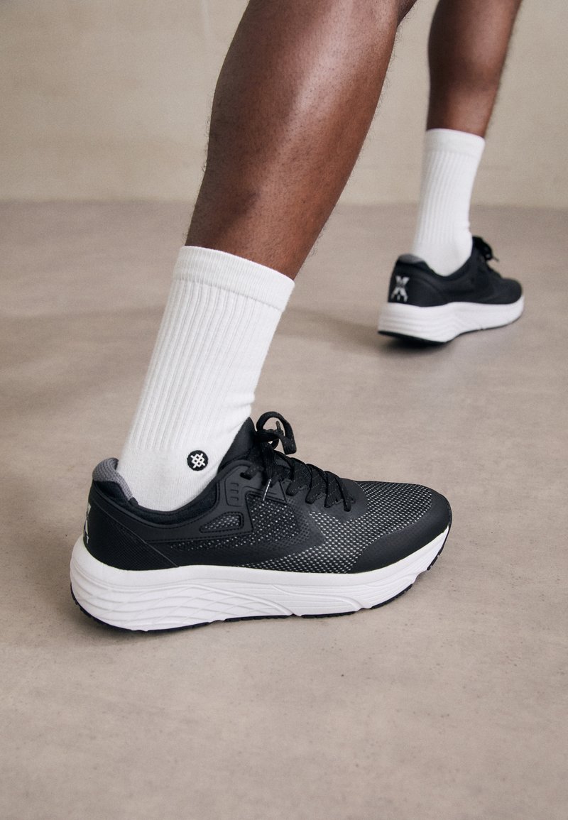 Schwarzer Sportschuh aus Netz- und glatten Materialien, mit weißer Sohle, grauen Akzenten und schwarzem Schnürverschluss. Getragen mit weißen gerippten Socken.