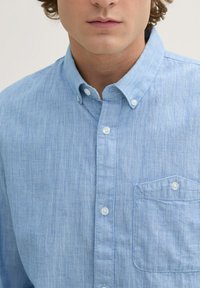 Hellblaues Hemd mit Knopfleiste aus strukturiertem Stoff, mit einer Brusttasche und weißen Knöpfen. Der Kragen liegt flach am Hals.