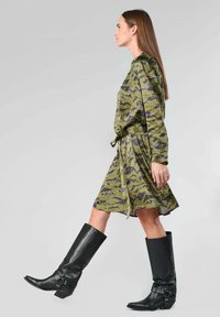 Grünes Camouflage-Kleid mit langen Ärmeln und Taillengürtel, kombiniert mit knielangen schwarzen Lederstiefeln mit einer Schnallen-Detail.