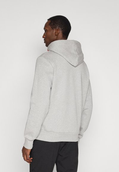 Mann, der nach links schaut, trägt eine hellgraue Kapuzenjacke und schwarze Hose vor weißem Hintergrund.