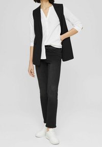 Gilet noir sur mesure porté sur une chemise blanche à boutons, associé à un jean noir et des baskets blanches ; coupe droite et détails minimalistes.