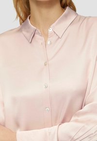 Blusa satinata rosa chiaro con colletto classico, bottoni simili alle perle e maniche lunghe con una texture liscia. Finitura lievemente lucida.