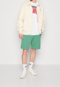 Krémová fleece bunda, bílý tričko s grafikou, zelené šortky a bílé tenisky se světlejšími akcenty. Model stojí s rukama v kapsách.
