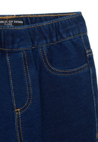 Jeans de mezclilla azul oscuro con cintura elástica y costura amarilla de contraste en los bolsillos y las costuras, etiquetados "República de Denim Flare."