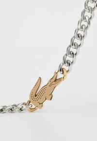 Bracelet en chaîne en métal argenté avec un charme crocodile en or. La chaîne présente des maillons entrelacés, tandis que le charme affiche un gravure détaillée.