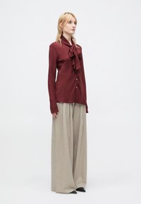 Bordeauxfarbene Bluse mit Schleifendetail, langen Ärmeln und Knopfverschluss; kombiniert mit hellgrauen, weit geschnittenen Hosen und schwarzen spitzen Schuhen.