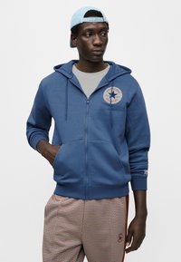 Μπλε φούτερ με φερμουάρ και κουκούλα με κορδόνι, διακοσμημένο με στρογγυλό λογότυπο. Φορεμένο πάνω από γκρι μπλουζάκι, σε συνδυασμό με καρό π αντελόνι.