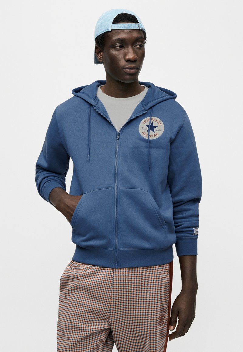 Blå hættetrøje med lynlås, snor i hætten og rund logo-detalje. Båret over en grå skjorte, kombineret med ternede bukser.